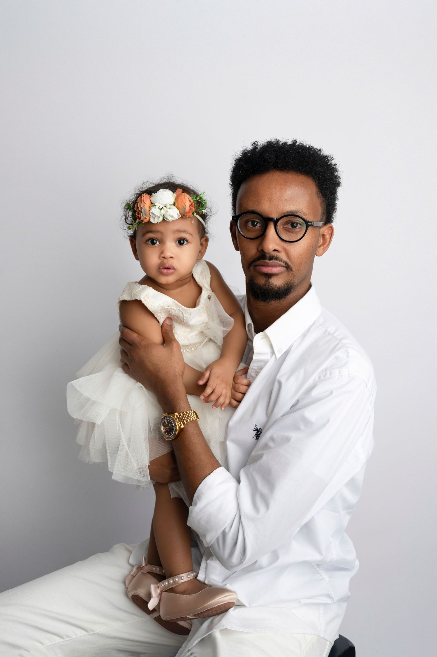 A man in a white shirt and glasses holds a baby girl in a white tutu dress with a floral headband.