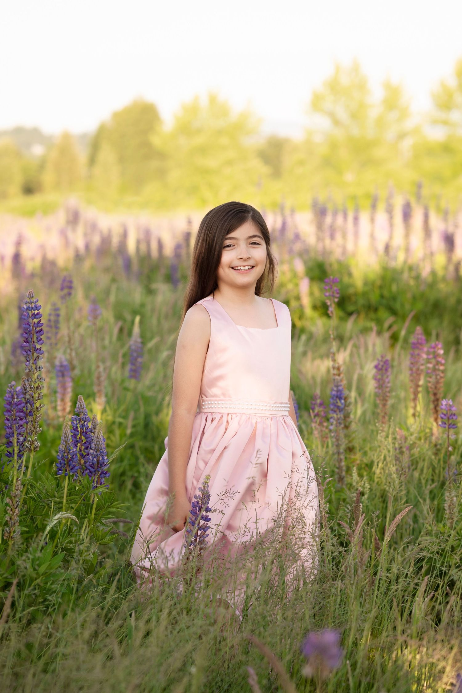 A smiling young girl in a pink dress standing in a field of purple lupine flowers on a sunny day.