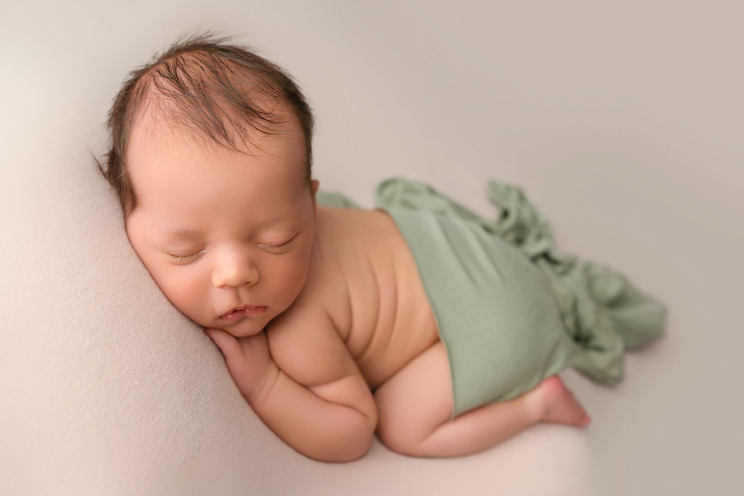 Peaceful sleeping newborn baby wearing mint green wrap on soft beige backdrop during professional photo session.