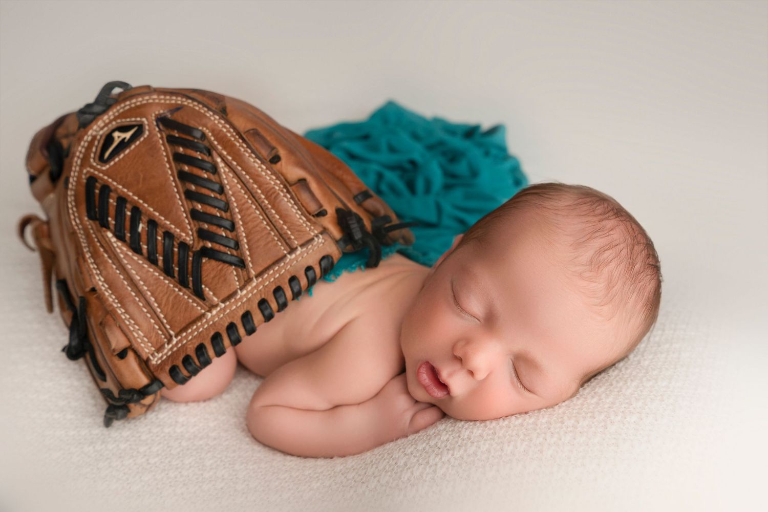 Sleeping newborn rests peacefully next to a brown baseball glove on white backdrop with teal accent.