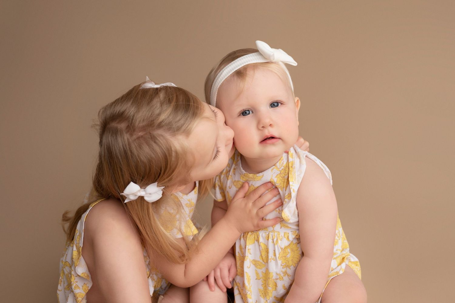 Two young children in matching yellow floral dresses share a sweet moment against a neutral backdrop.