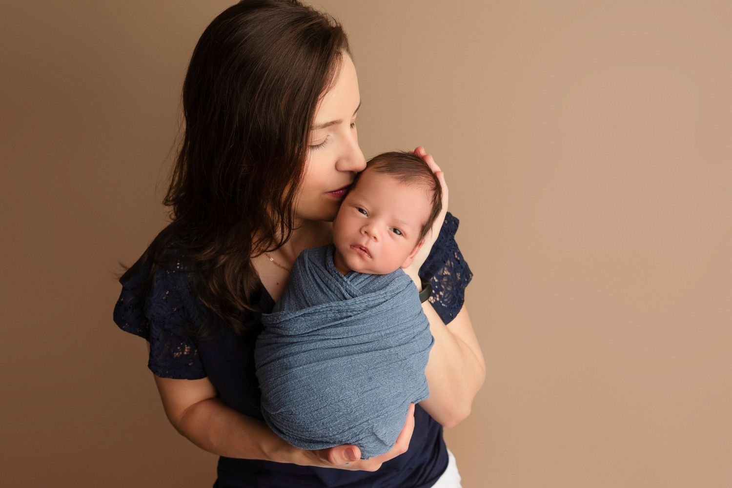 A parent wearing a dark top holds a baby close in a blue wrap carrier against a neutral beige background.