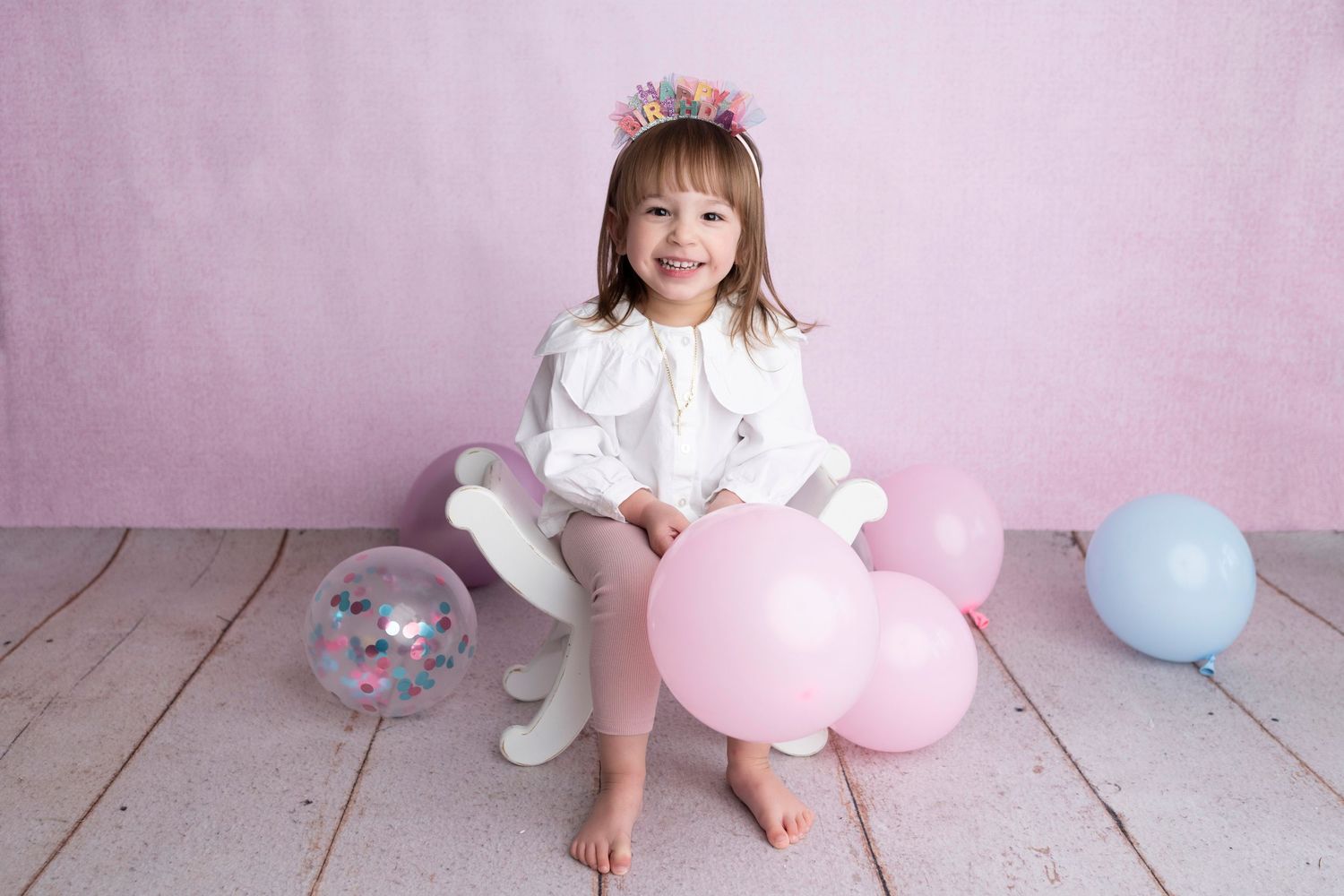 A smiling little girl in a white dress and birthday crown sits on a toy horse surrounded by colorful balloons on a pink backdrop.