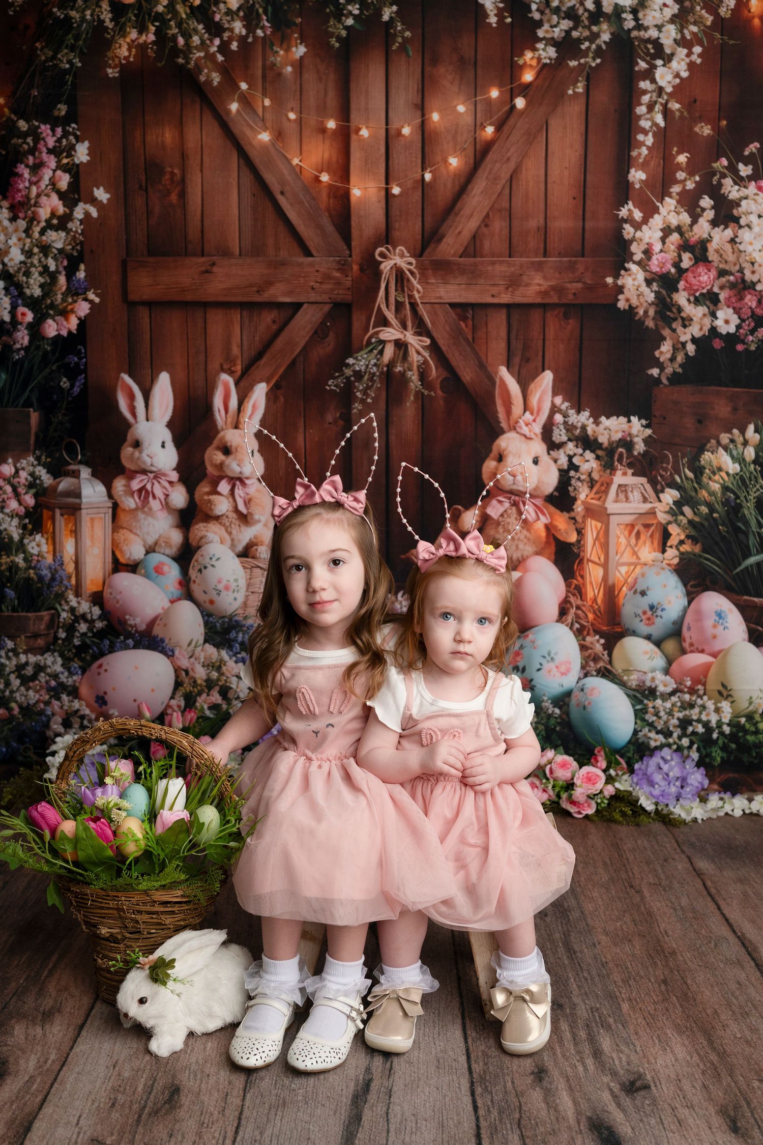 Two little girls in pink dresses pose with Easter eggs, bunnies, and spring flowers in a charming barn-themed holiday photo.