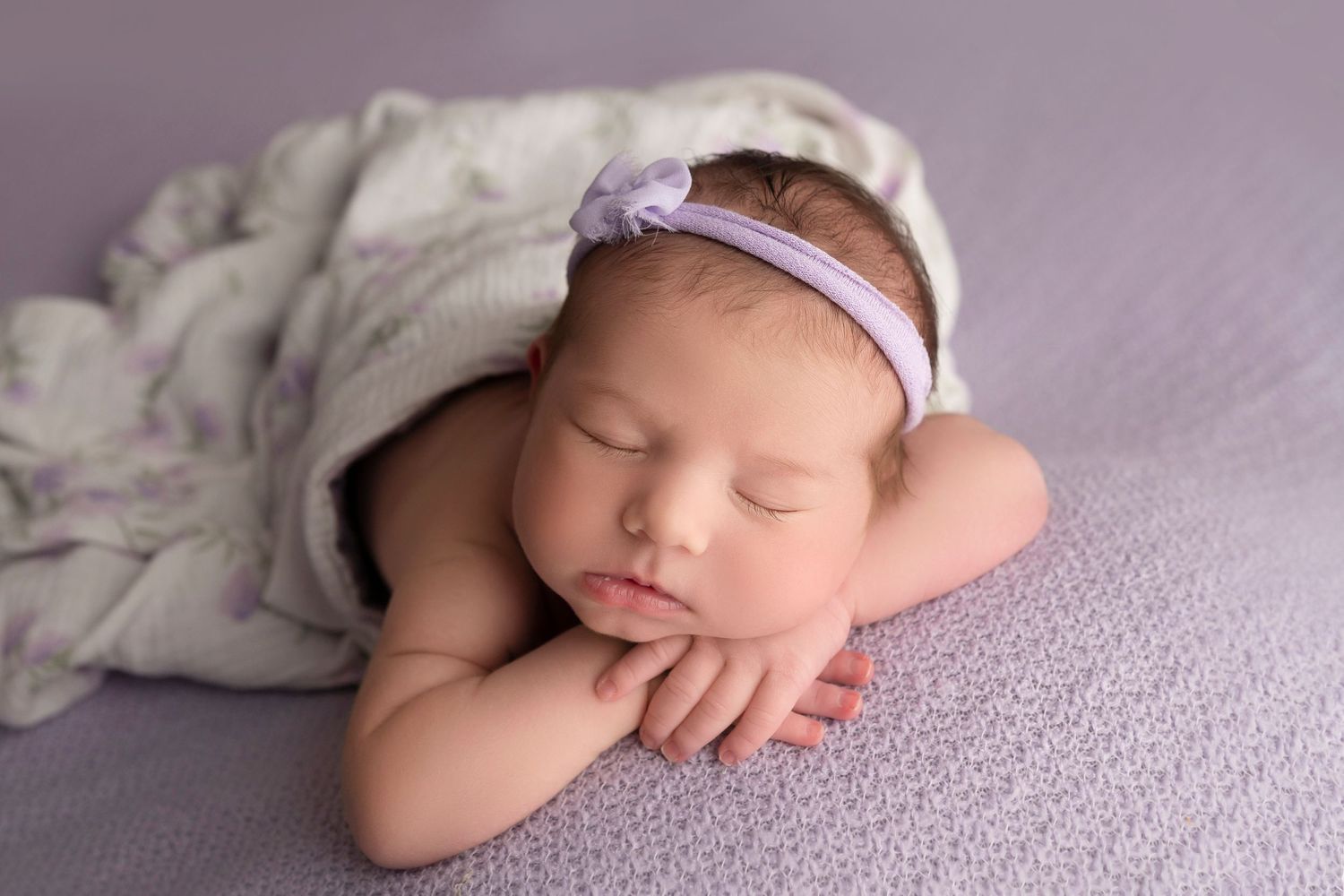 Peaceful newborn baby sleeping with hands folded under chin, wearing lavender headband, wrapped in soft purple blanket.