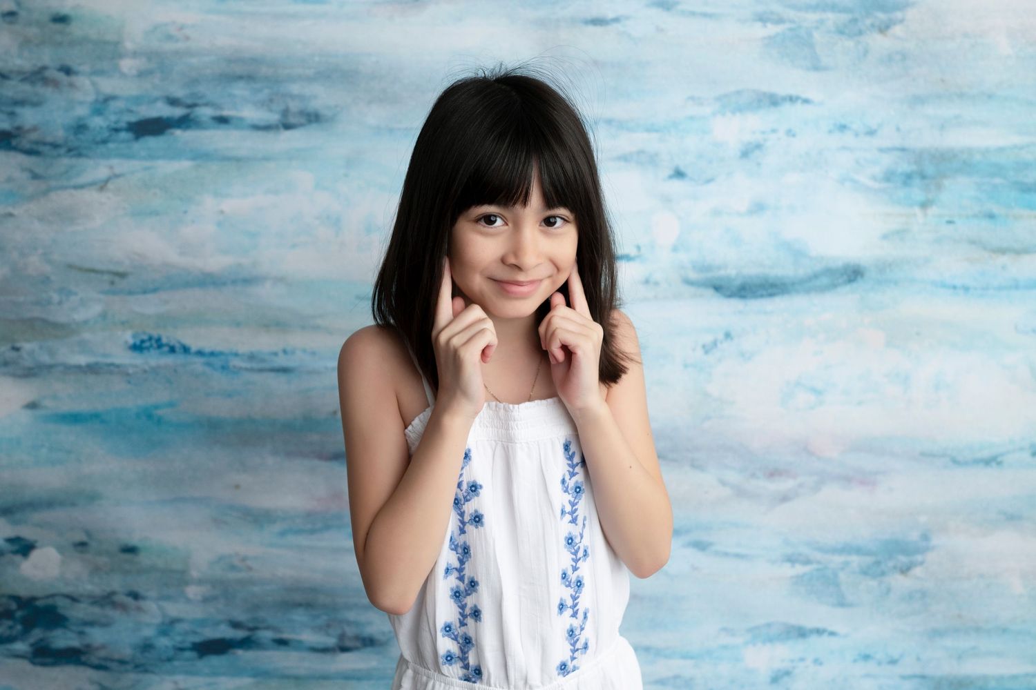 Young girl in white dress posing against blue watercolor backdrop, smiling with hands near face.