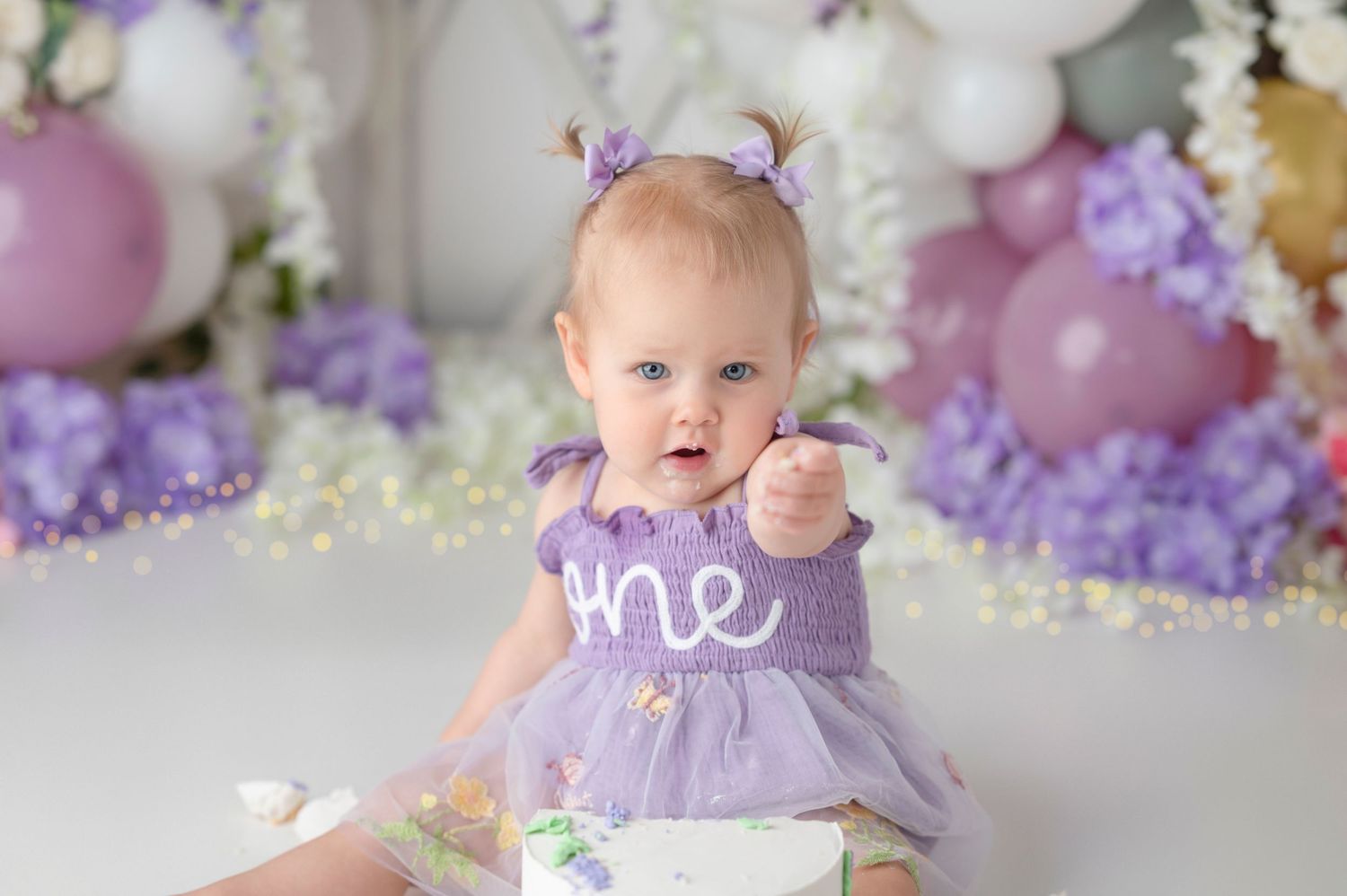Blue-eyed baby girl in purple tutu dress reaches toward camera beside smash cake at first birthday.