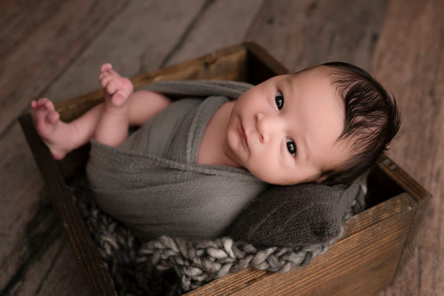 Newborn baby swaddled in gray wrap, resting in a rustic wooden box with knit blanket, looking up with bright eyes.