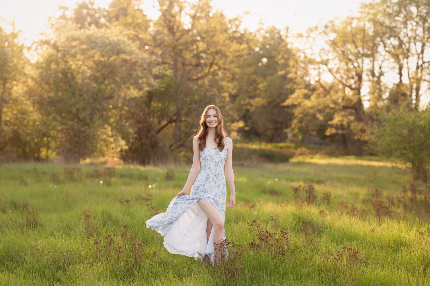 Young woman in flowing floral dress walks through sunlit green meadow with golden trees in background.