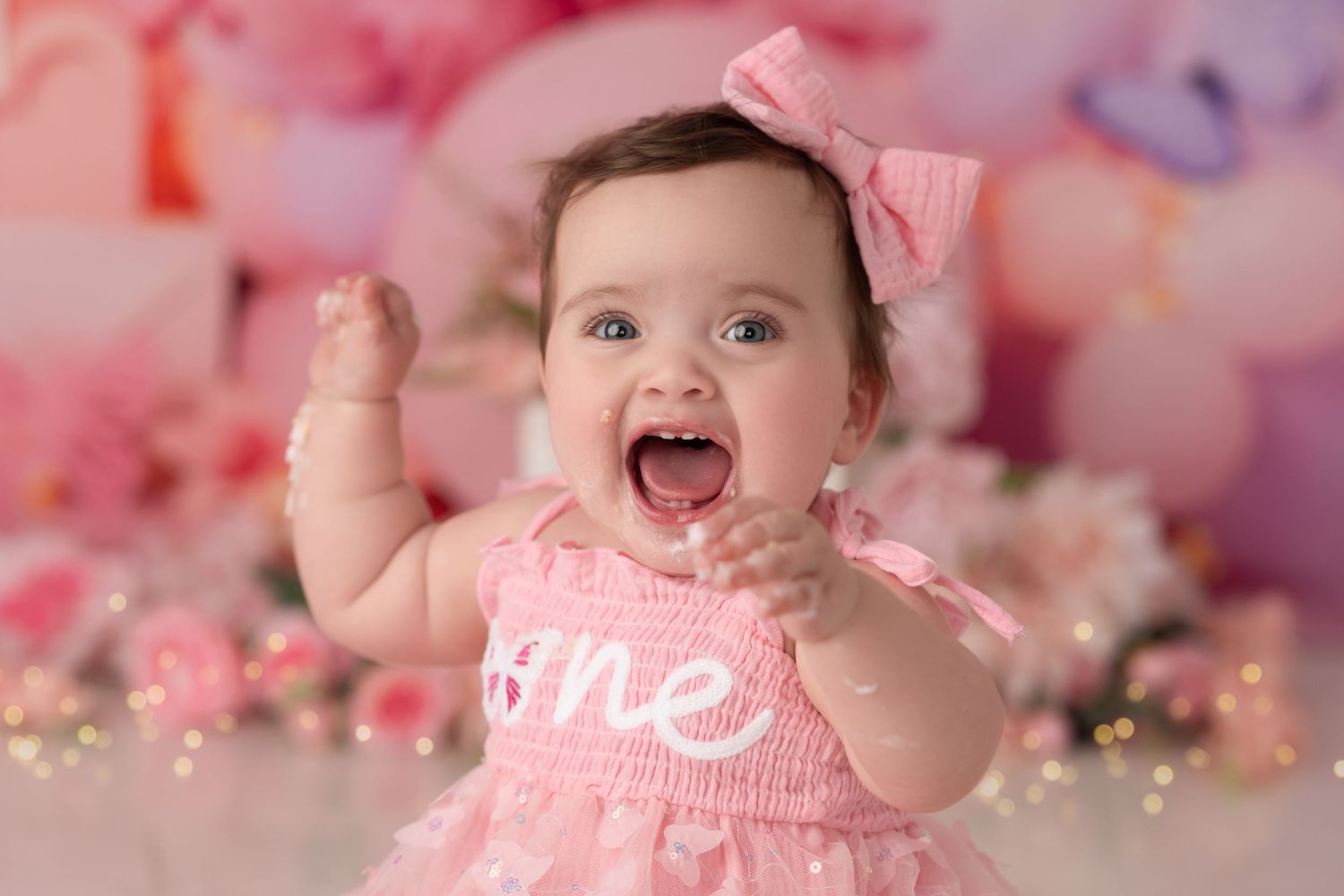A joyful baby girl in a pink dress with a bow celebrates her first birthday against a soft floral backdrop.