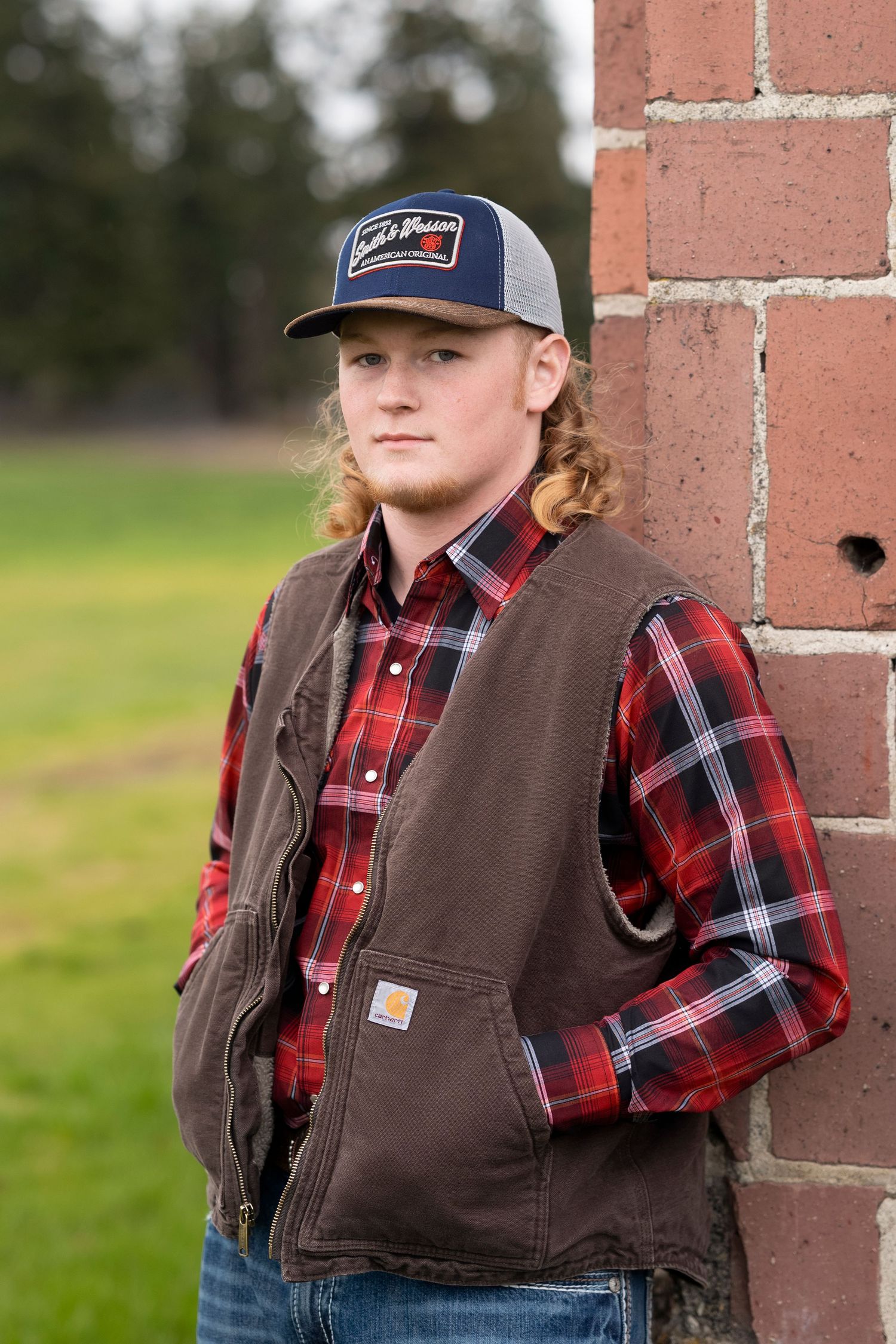 Person wearing a brown Carhartt vest and red plaid shirt poses against brick wall in outdoor setting.