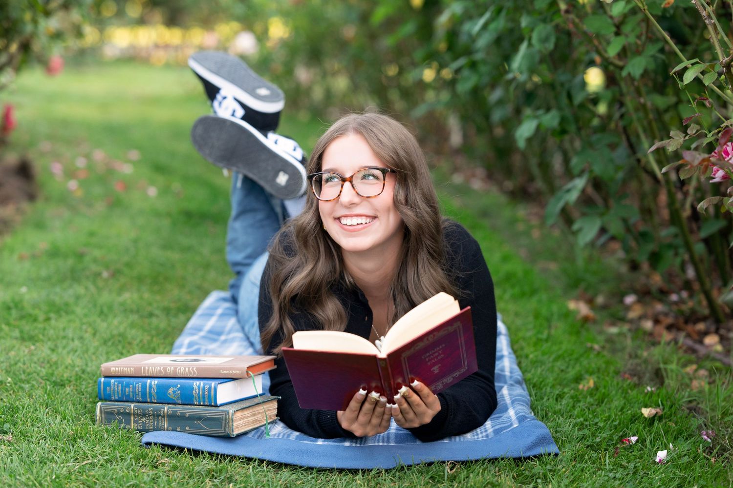 A person lies on a blanket reading a book outdoors on a sunny day surrounded by green grass and trees.