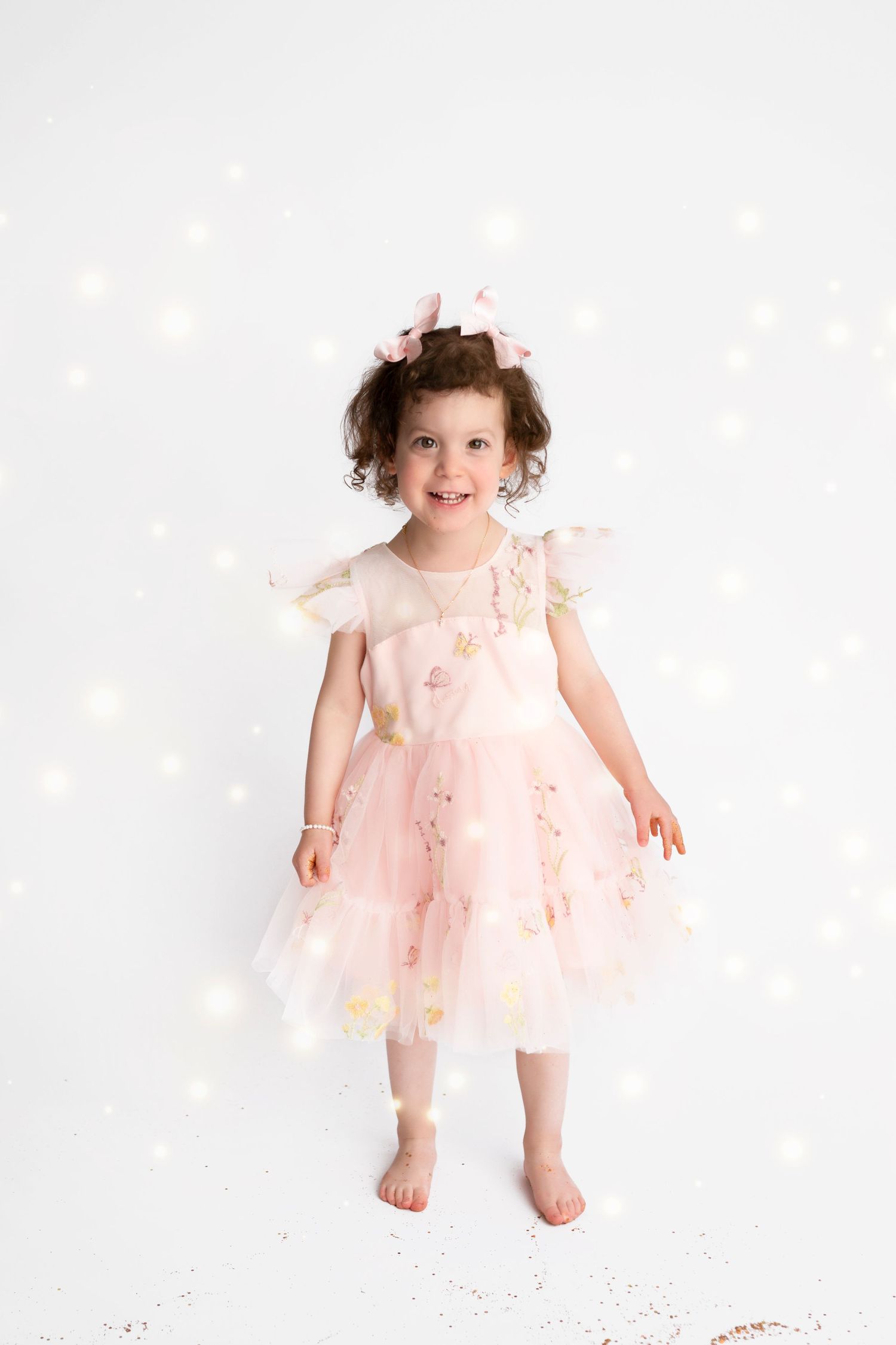 A toddler wearing a pink tulle dress twirls and plays with sparkles on a white background.