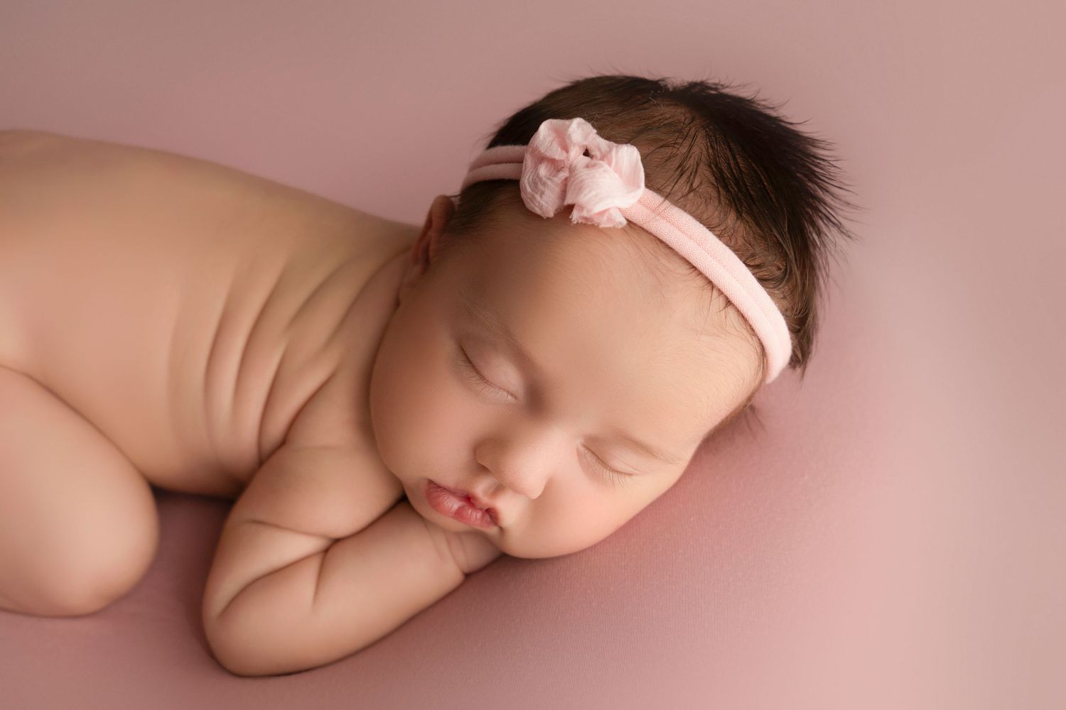 Peaceful newborn portrait with pink headband resting on pink background.