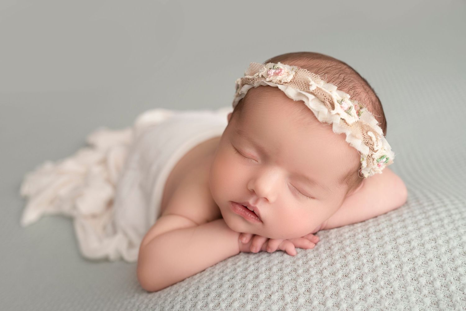 Sleeping newborn wearing delicate lace headband rests on soft gray textured blanket.