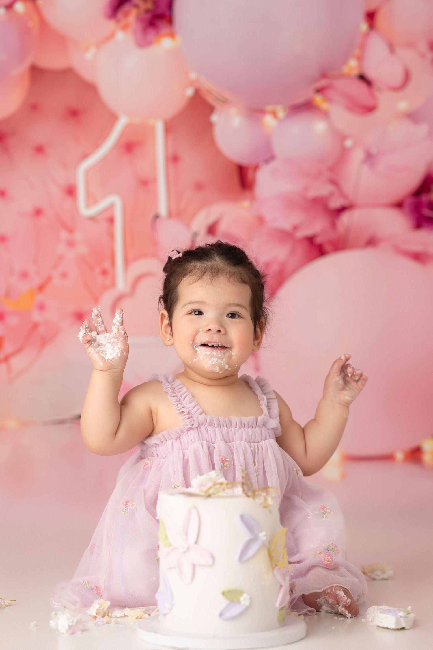 Pink balloon backdrop with a small child in a white dress celebrating with cake and frosting.