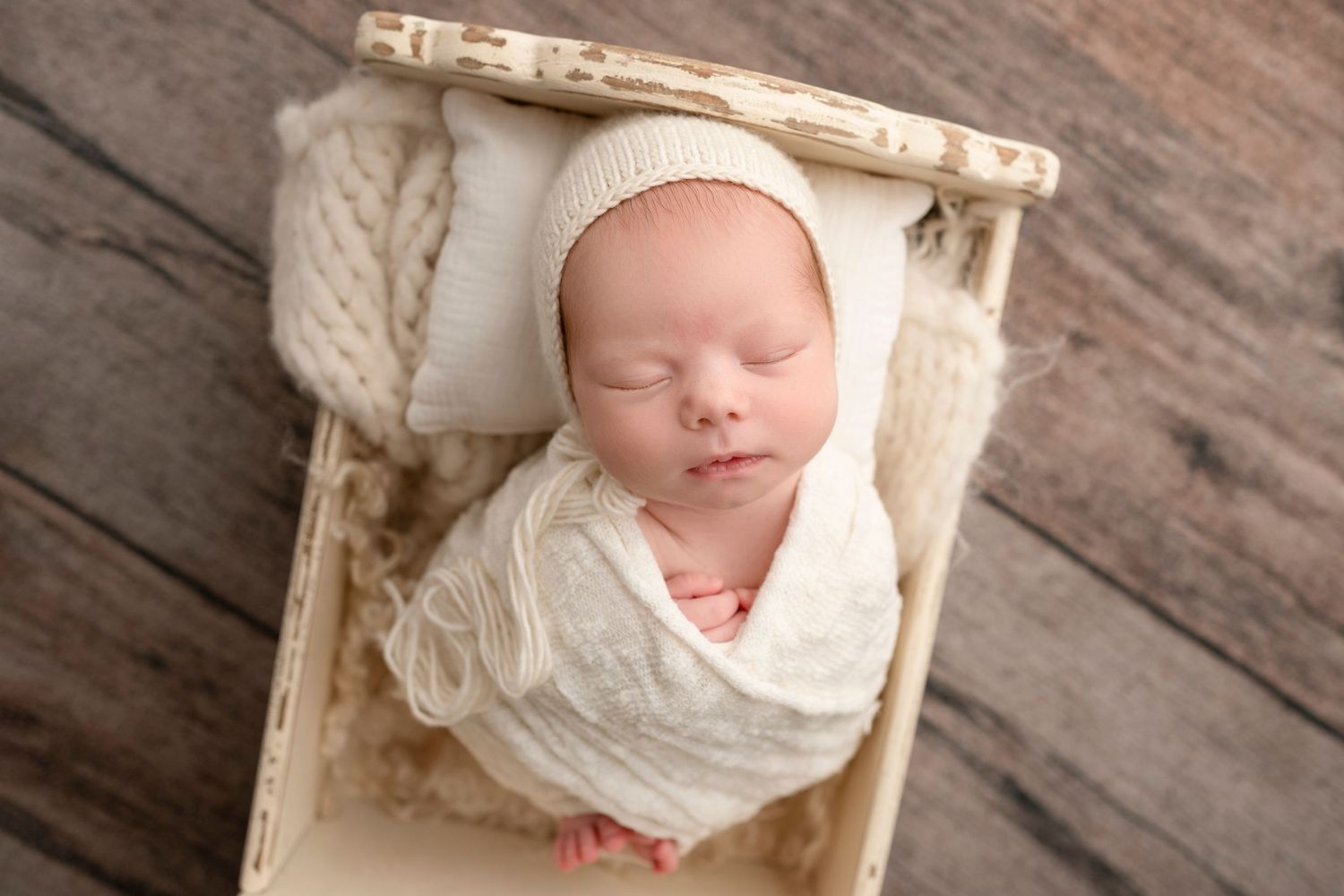 Sleeping newborn baby wrapped in cream knit swaddle wearing bonnet, posed in small wooden bed on rustic wood backdrop.