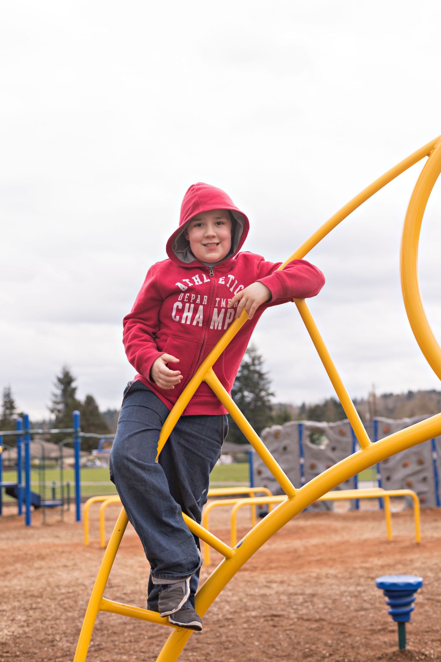 A smiling child in a red hoodie climbs yellow playground equipment outdoors on a cloudy day.
