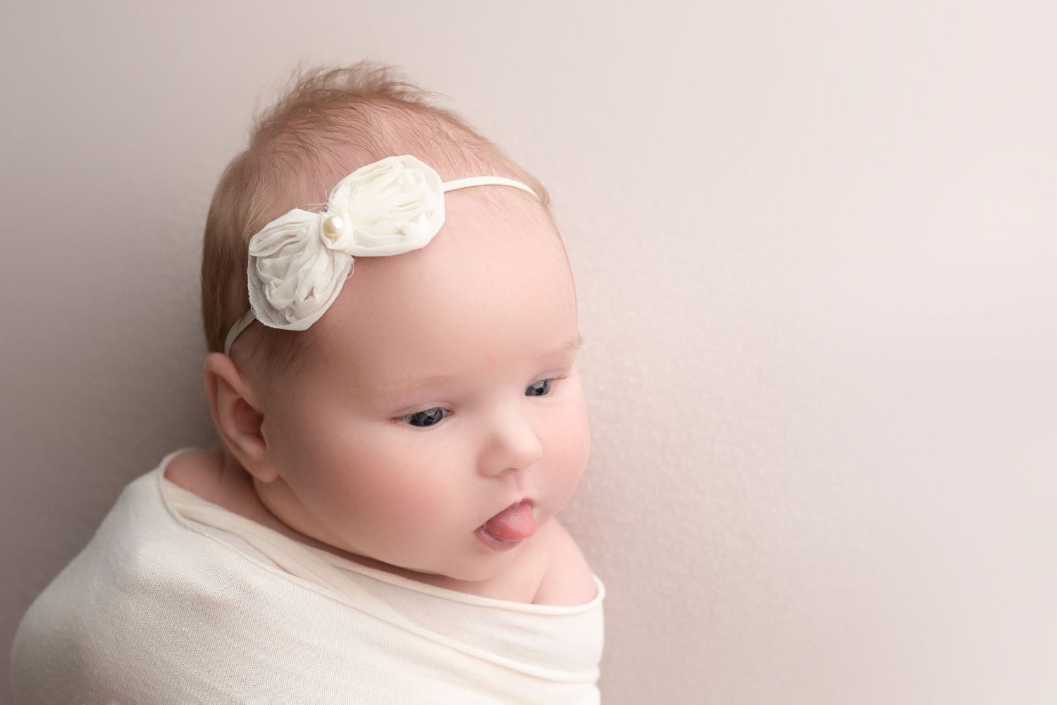 Newborn baby wrapped in white swaddle cloth wearing a delicate flower headband, posed against a soft neutral background.