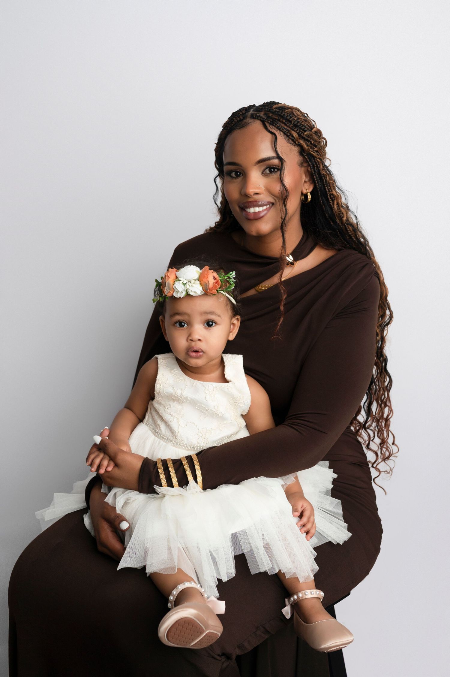 A woman in a brown dress holds a baby girl in a white tutu dress with a floral headband in a studio portrait.