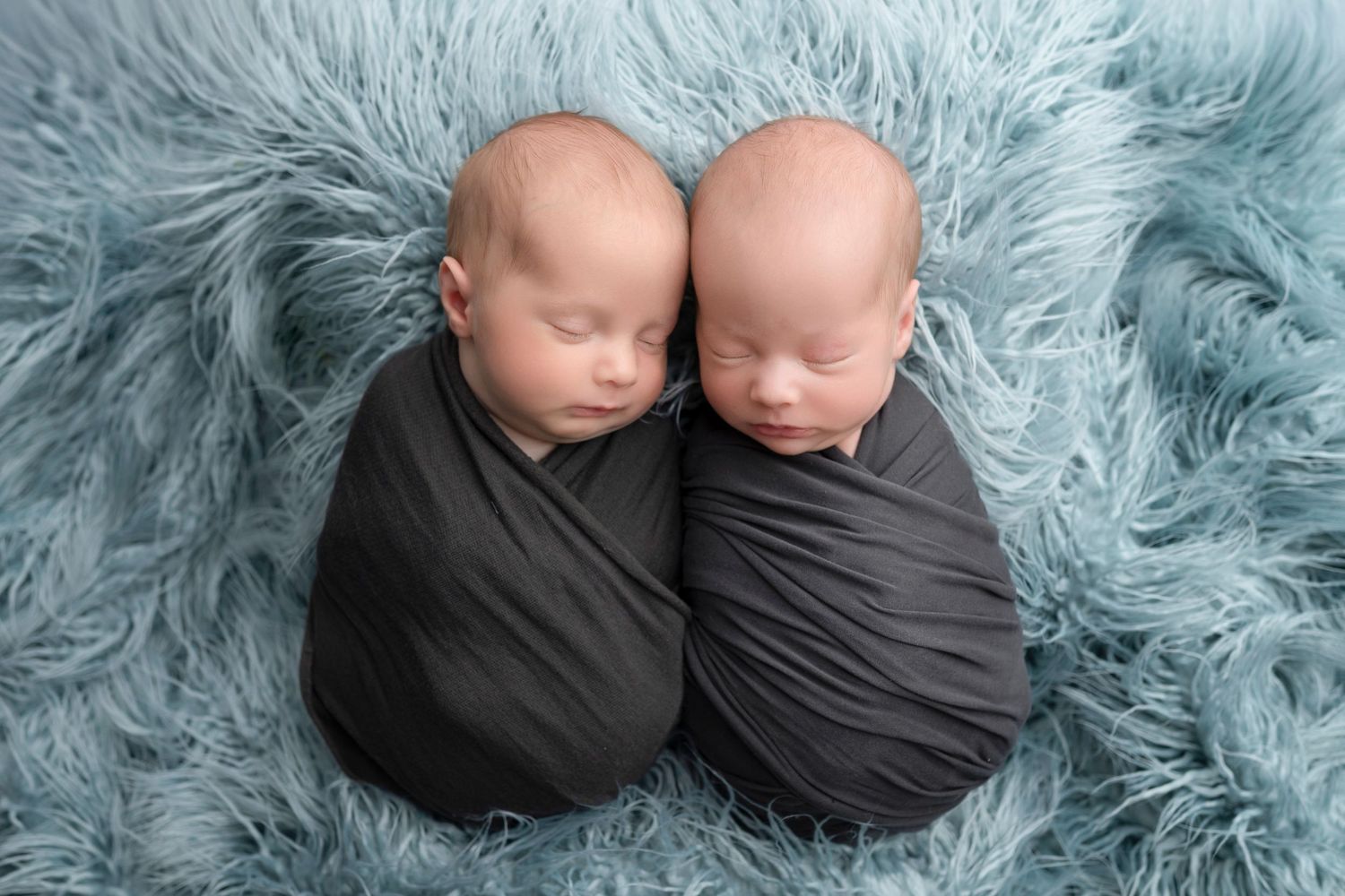 Two sleeping newborn twins swaddled in gray wraps lying together on a soft blue fluffy blanket.