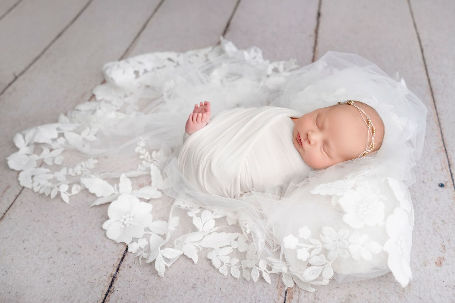 Newborn baby swaddled in white wrap sleeping on white floral lace blanket with delicate gold headband.