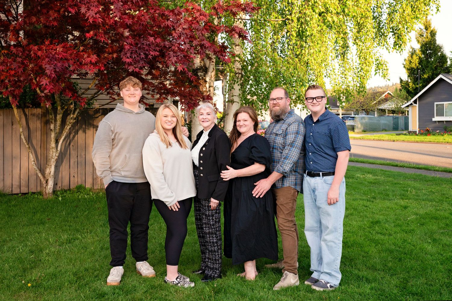 Six family members smiling together outdoors on green lawn with red autumn tree and birch tree background.