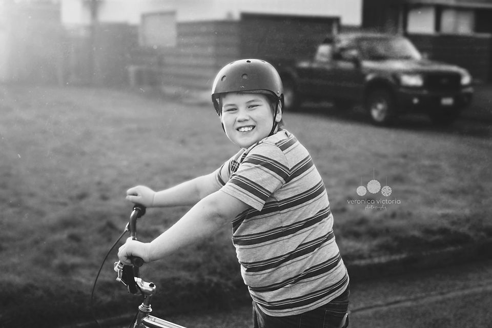 A smiling boy wearing a helmet rides a bike outdoors in a black and white photo.
