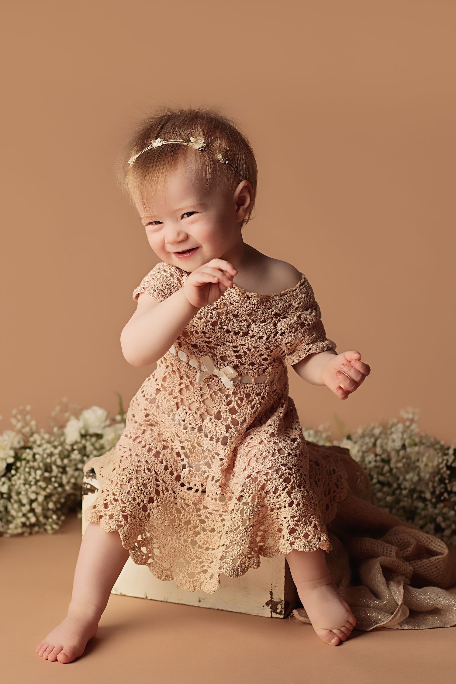 A baby wearing a delicate beige lace dress sits on a brown backdrop with baby's breath flowers as decoration.