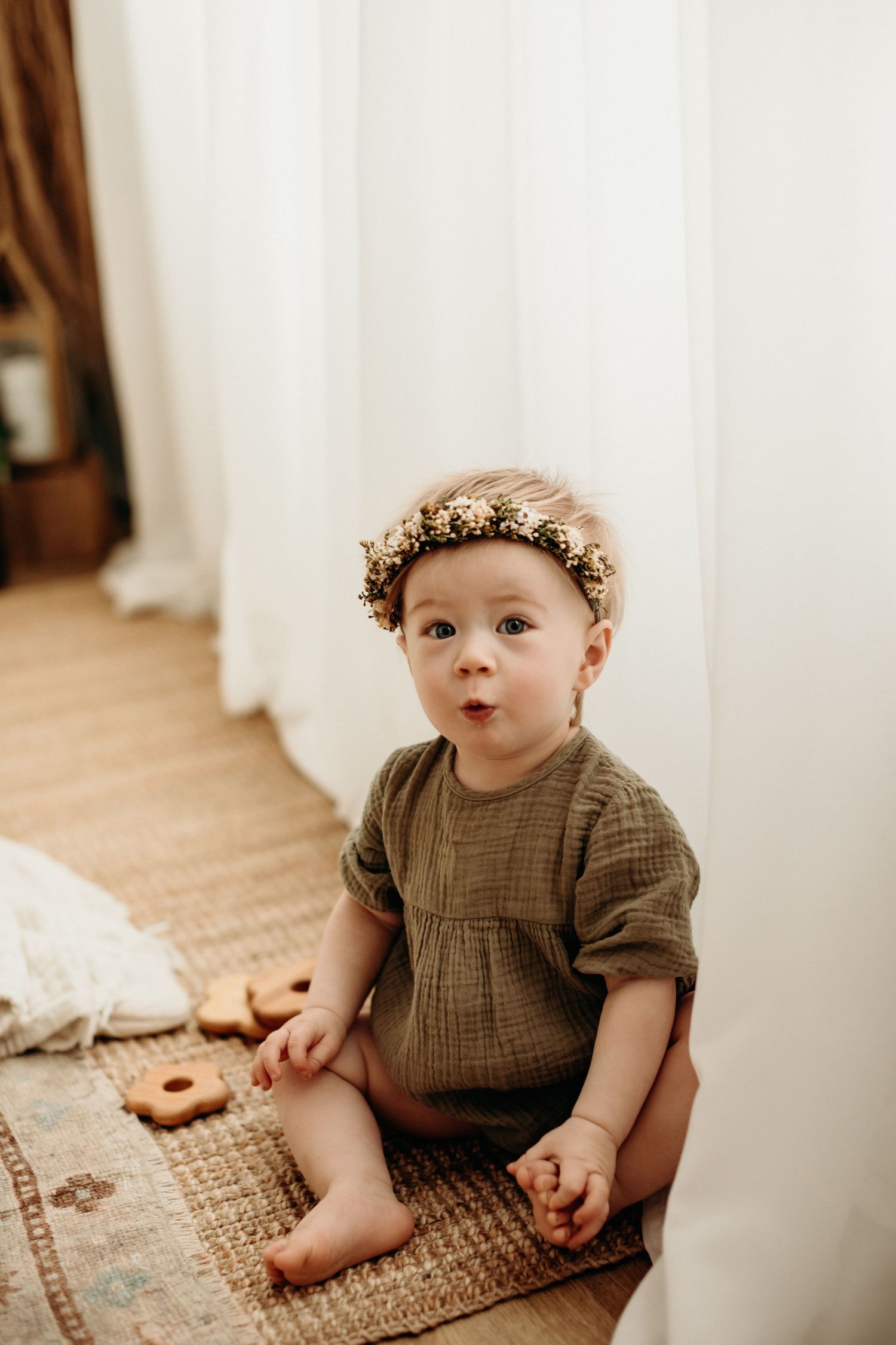 Baby wearing brown knit sweater and floral crown sits on patterned rug in minimalist indoor setting.