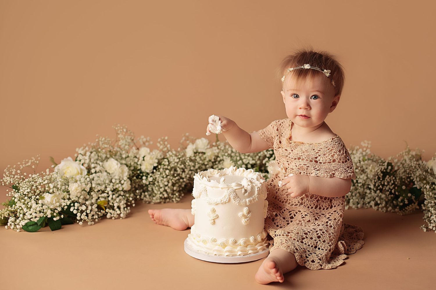 Baby wearing neutral colors sits next to floral decorations and small white cake during photo session in tan studio.
