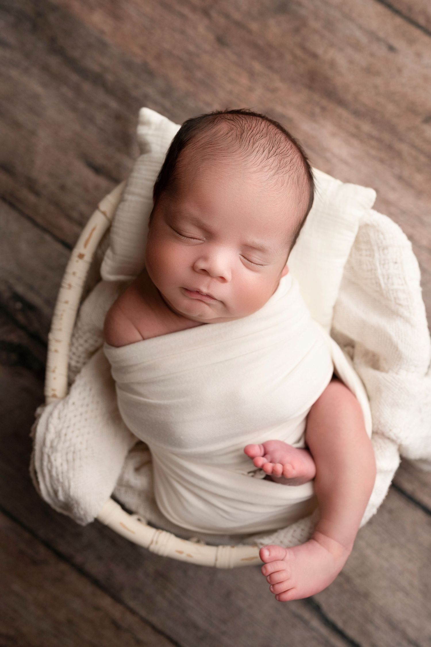 A sleeping newborn baby wrapped in soft white fabric rests peacefully on rustic wooden flooring.