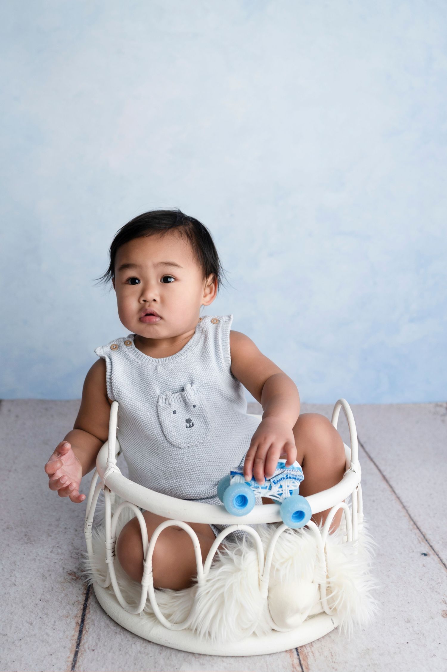 Baby sitting in white wicker basket with blue toy, wearing gray romper, against light blue background.