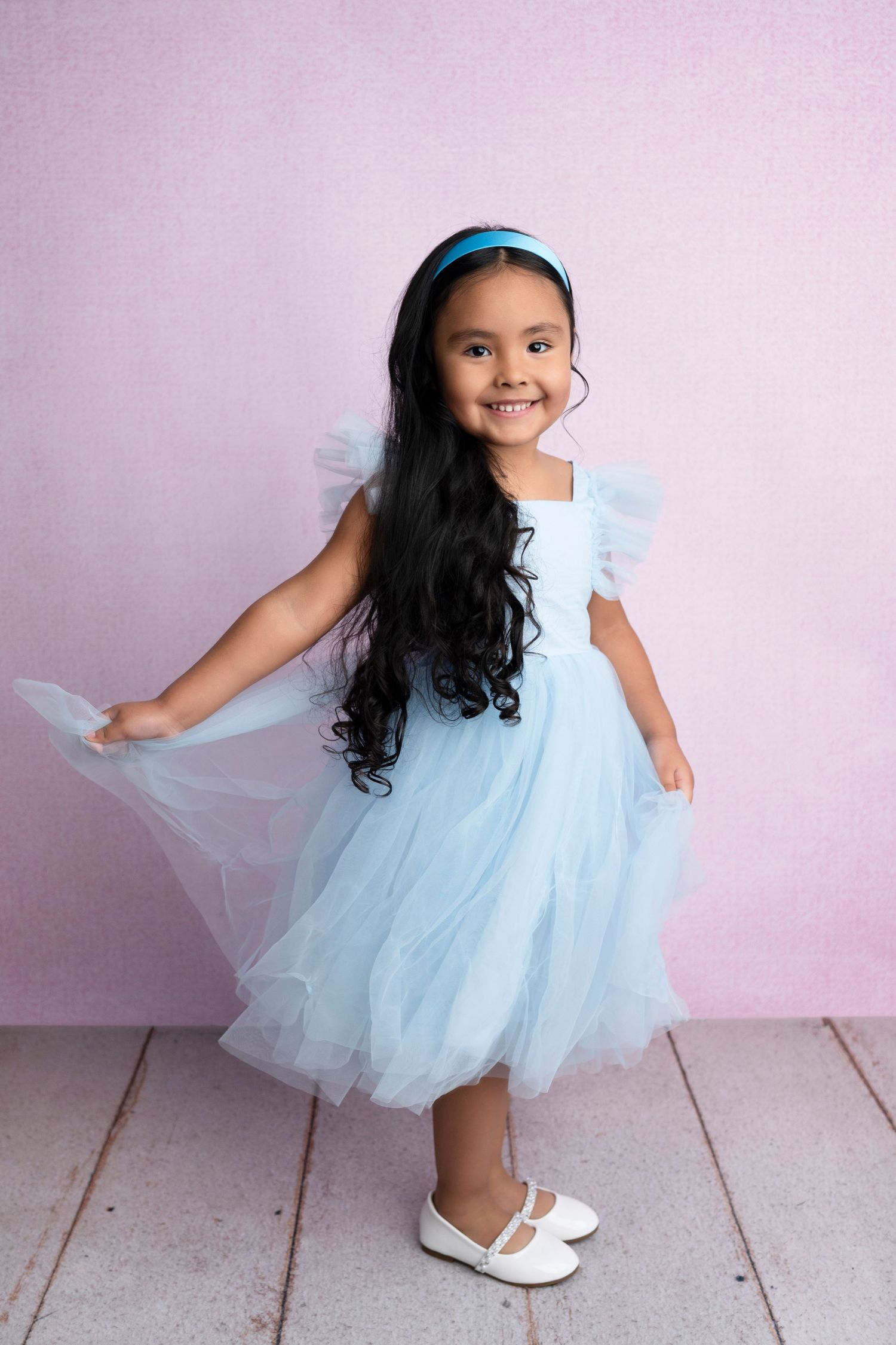 Young girl in light blue tulle dress with flutter sleeves, blue headband, and white shoes on pink background.