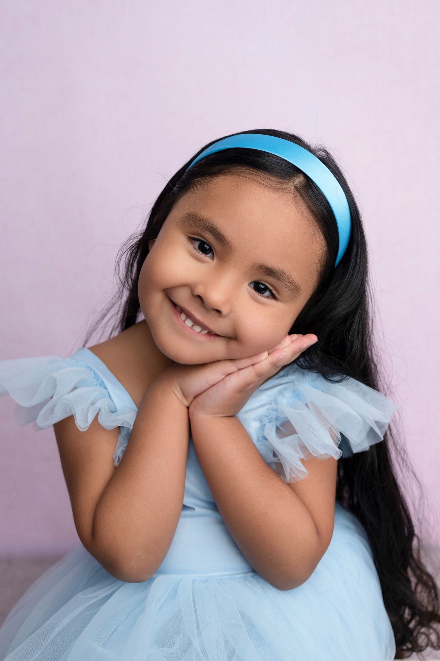 A smiling young girl in a blue tulle dress and headband poses with hands under chin against a pink background.