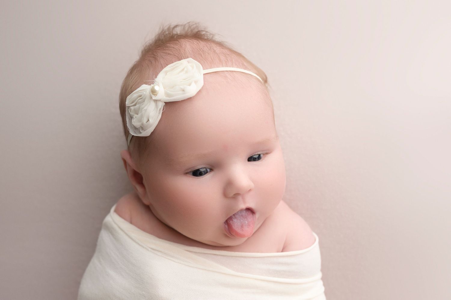 Adorable newborn baby wrapped in white, wearing a bow headband, sticking out tongue during professional photo session.