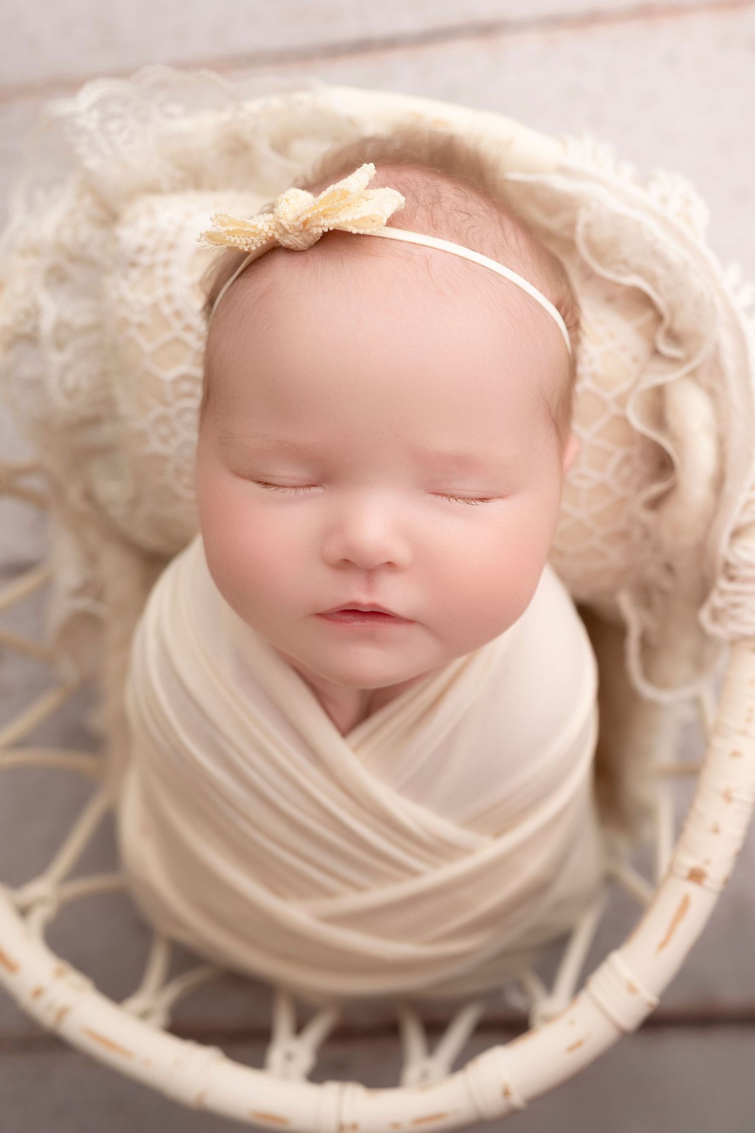 Sleeping newborn baby swaddled in cream wrap with bow headband, nestled in a white wicker basket.