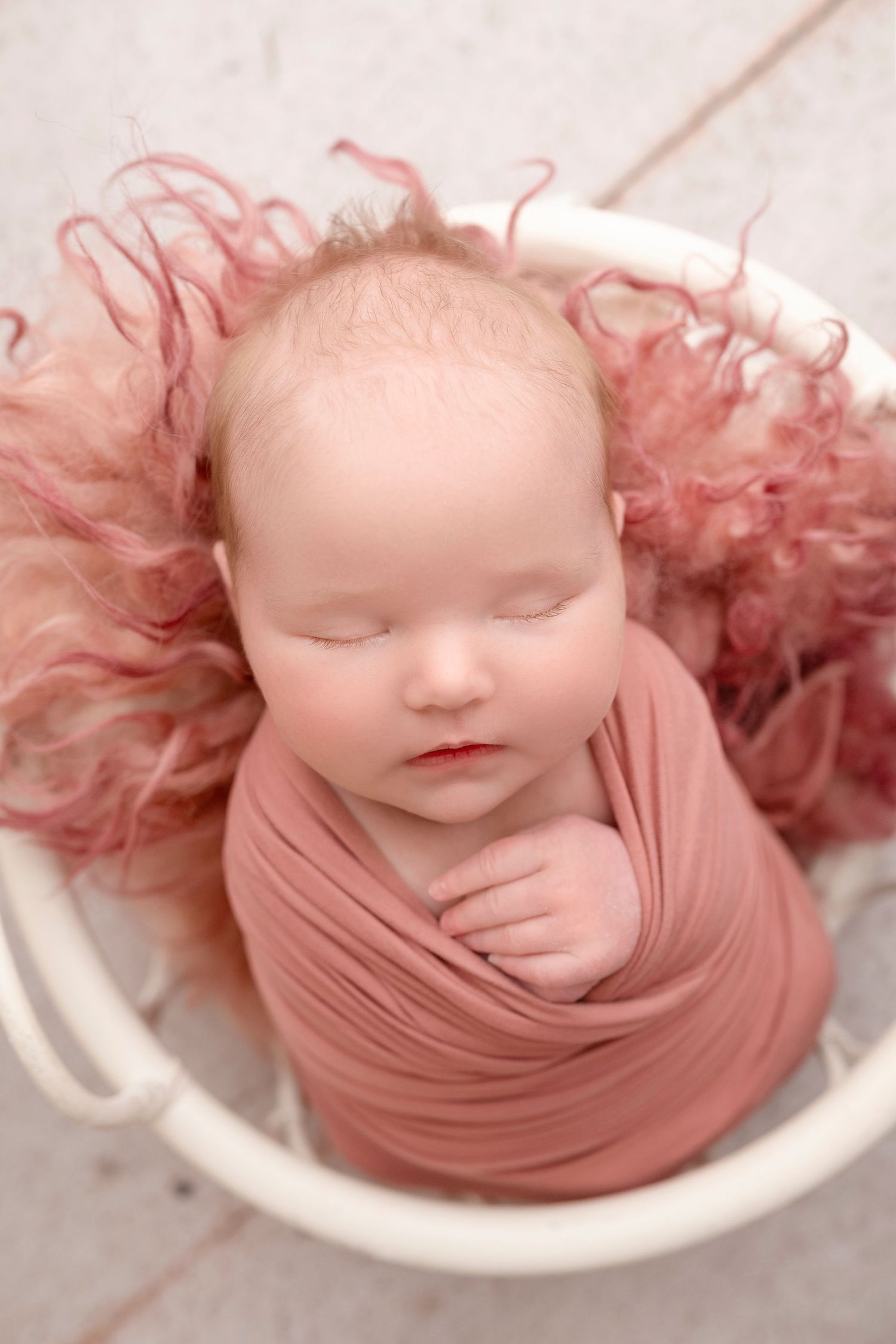 Sleeping newborn baby swaddled in pink wrap nestled in white bucket with fluffy pink fur, professional newborn photography.
