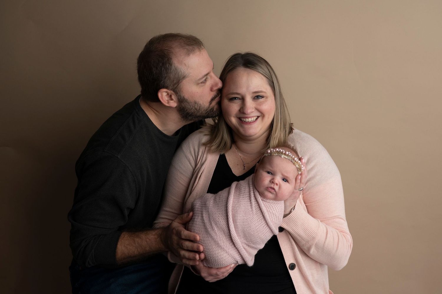A happy couple poses with their newborn baby, the man kissing the woman's cheek as she holds the infant.