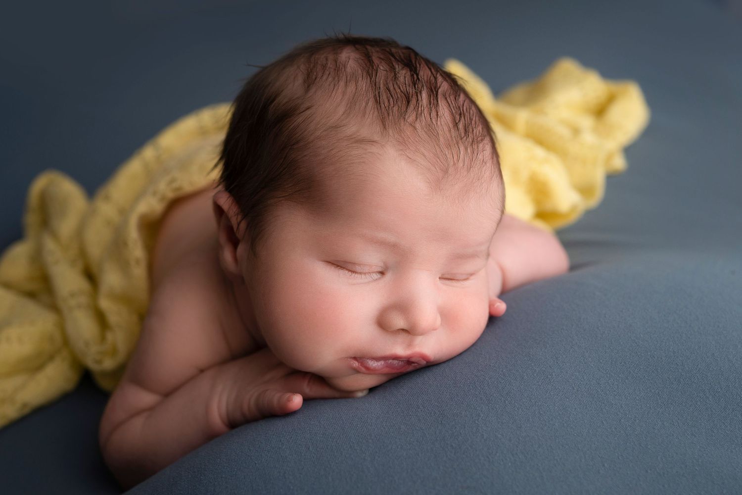 Sleeping newborn baby resting on blue backdrop with yellow knit wrap, peaceful close-up portrait photography.