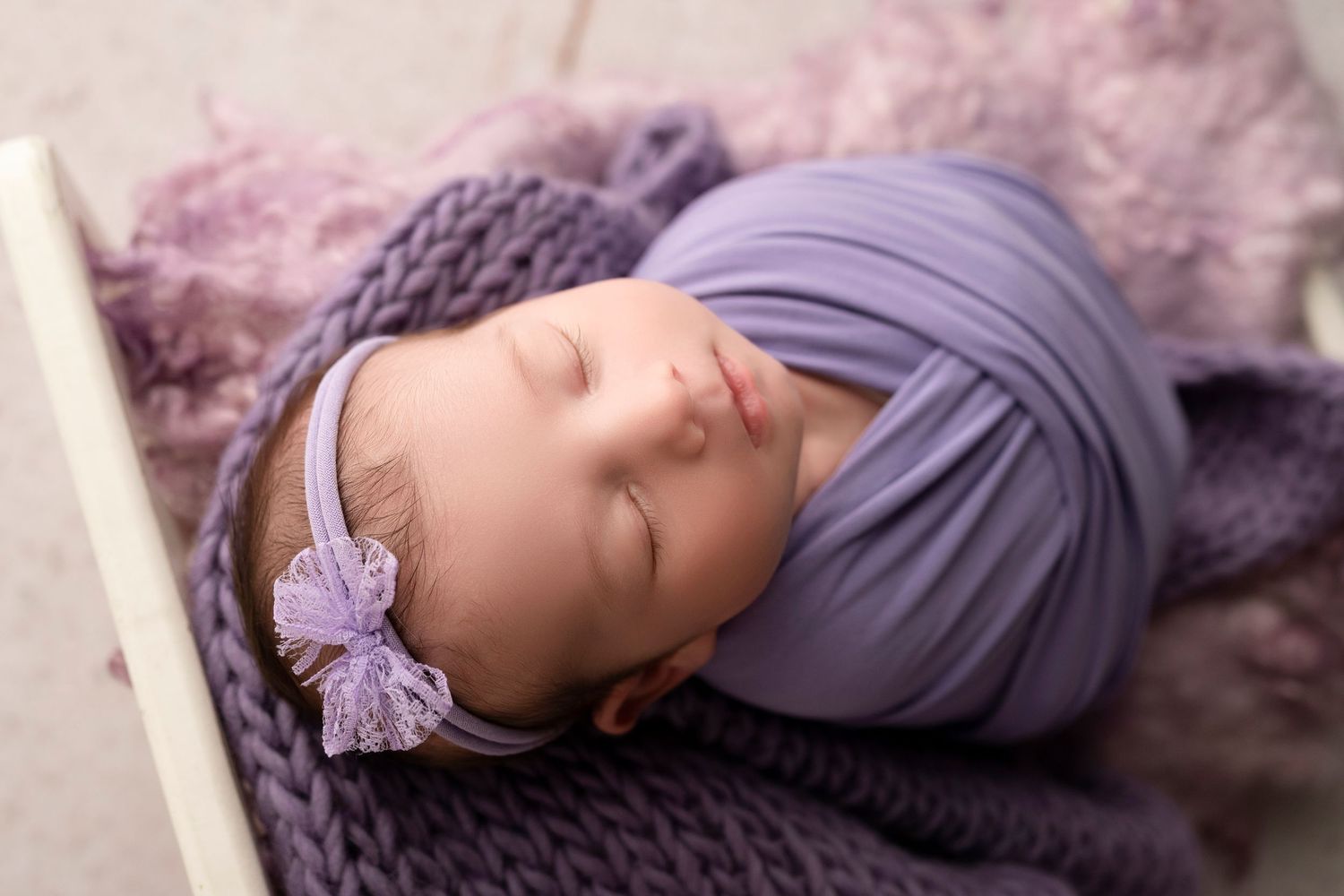 Newborn baby girl sleeping peacefully wrapped in purple swaddle with lavender bow headband in cozy photo session.