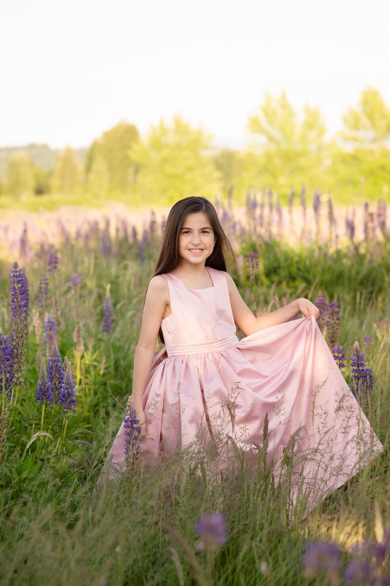 A smiling girl in a pink floral gown poses in a field of purple lupine flowers during golden hour.