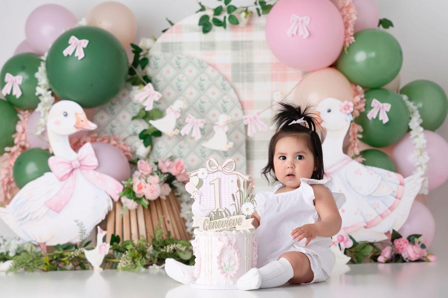 Baby girl in white dress sits by first birthday cake surrounded by pink and green balloons and white swan decorations.