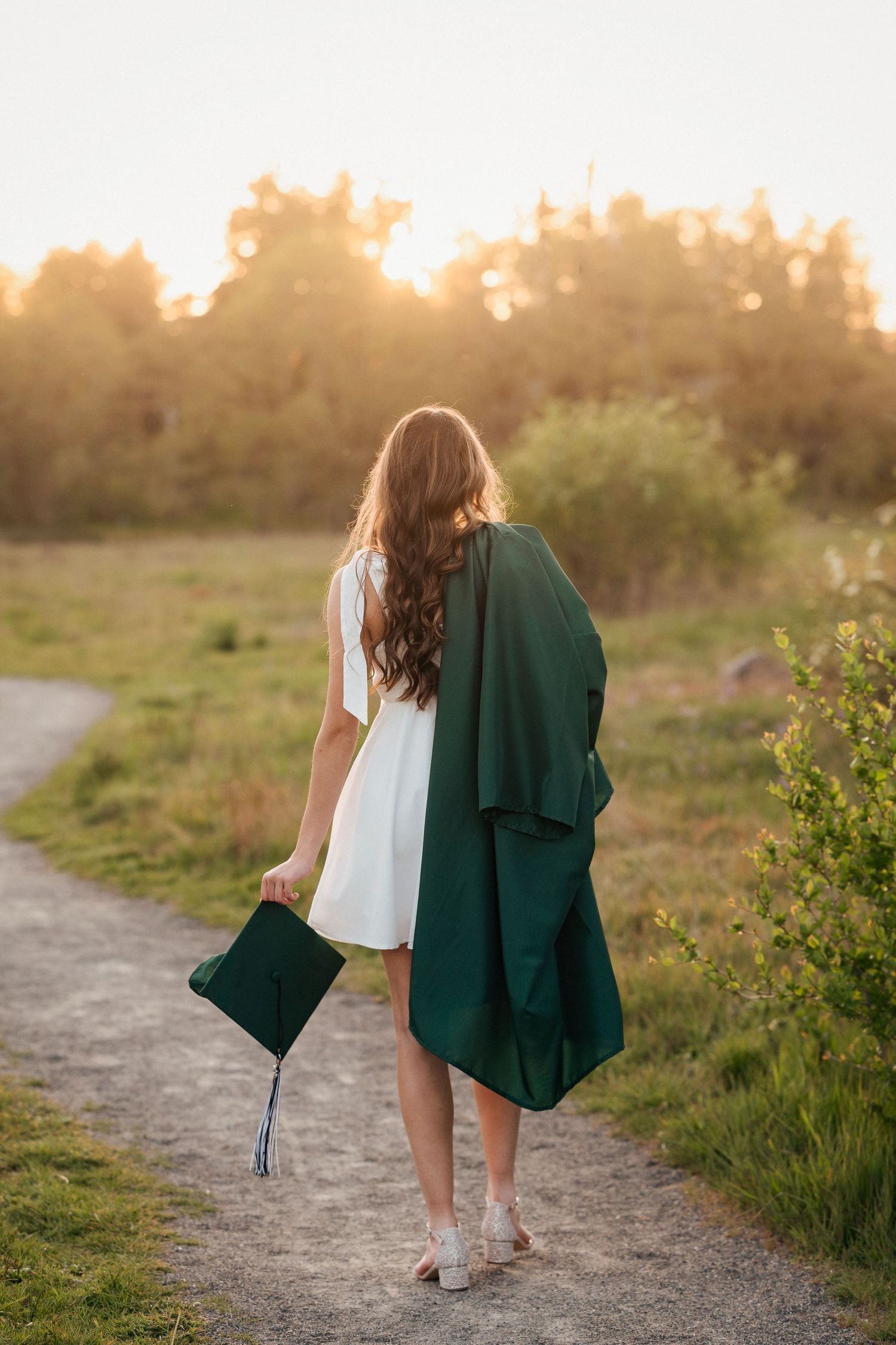 A person in a white dress and green graduation gown walks along a sunset-lit path through a grassy field.