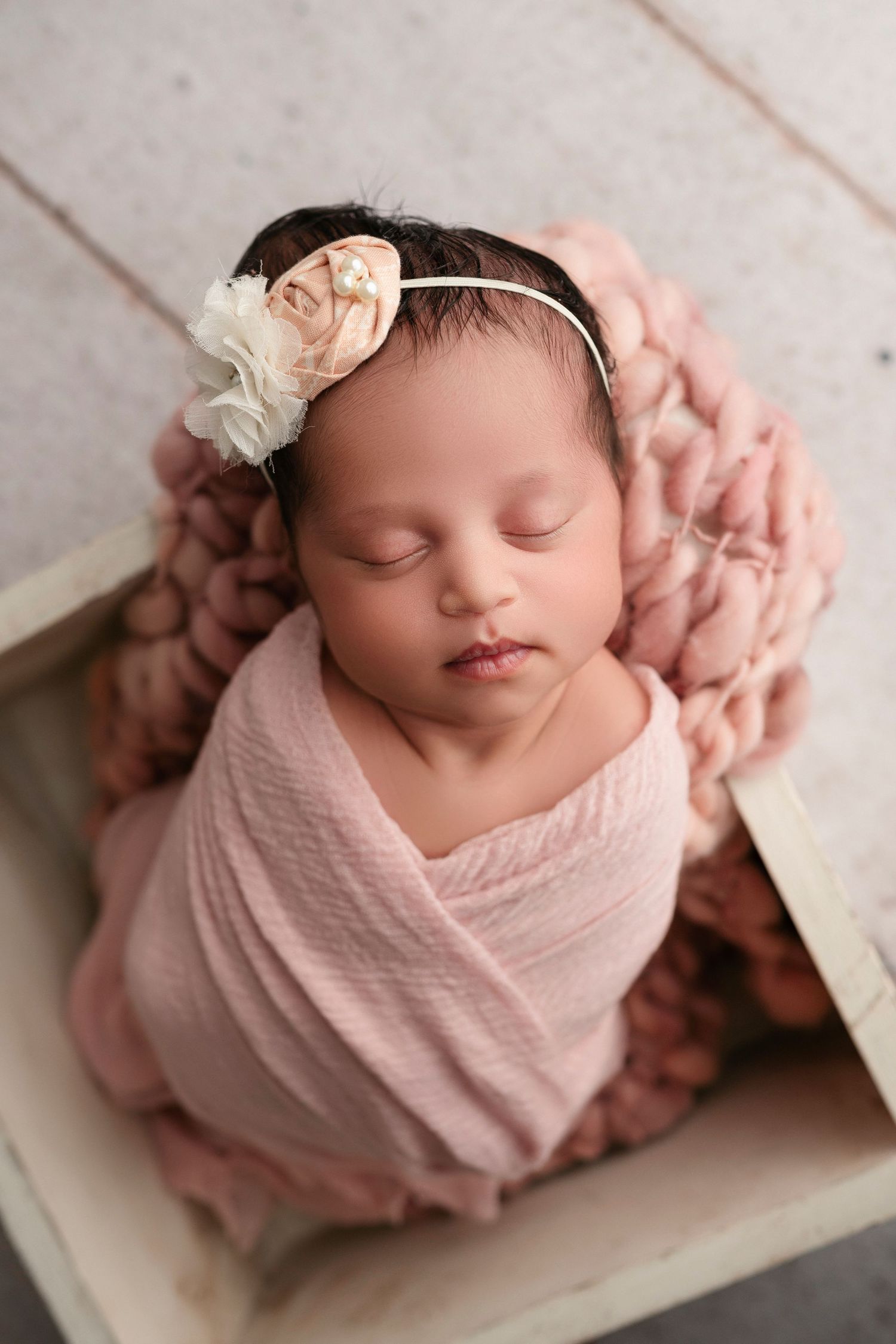 Sleeping newborn baby swaddled in soft pink fabric with delicate floral headband lying on white surface.