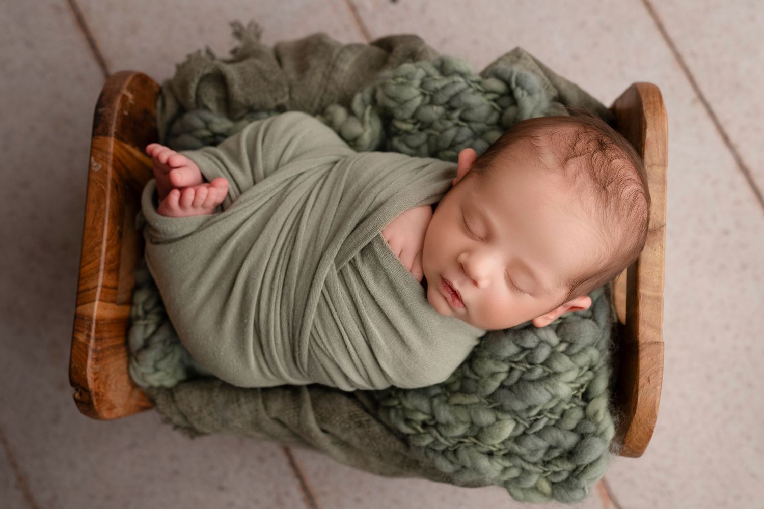 Baby resting in vintage wooden cradle with olive green swaddle and textured bedding.