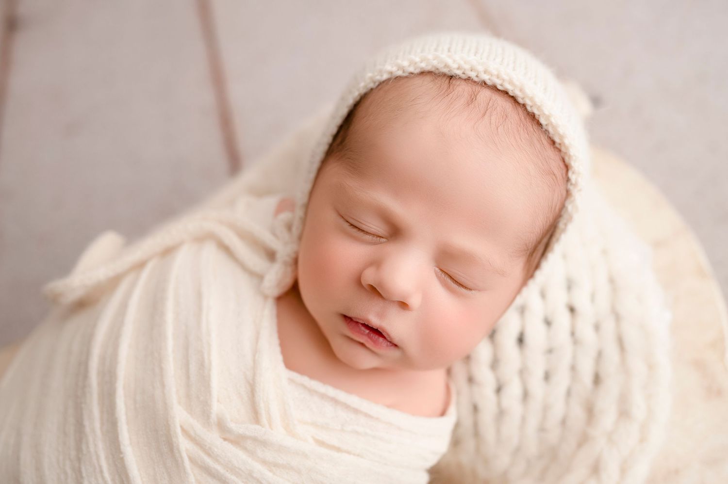 Peaceful newborn in cream wrap and knit bonnet against soft neutral background.