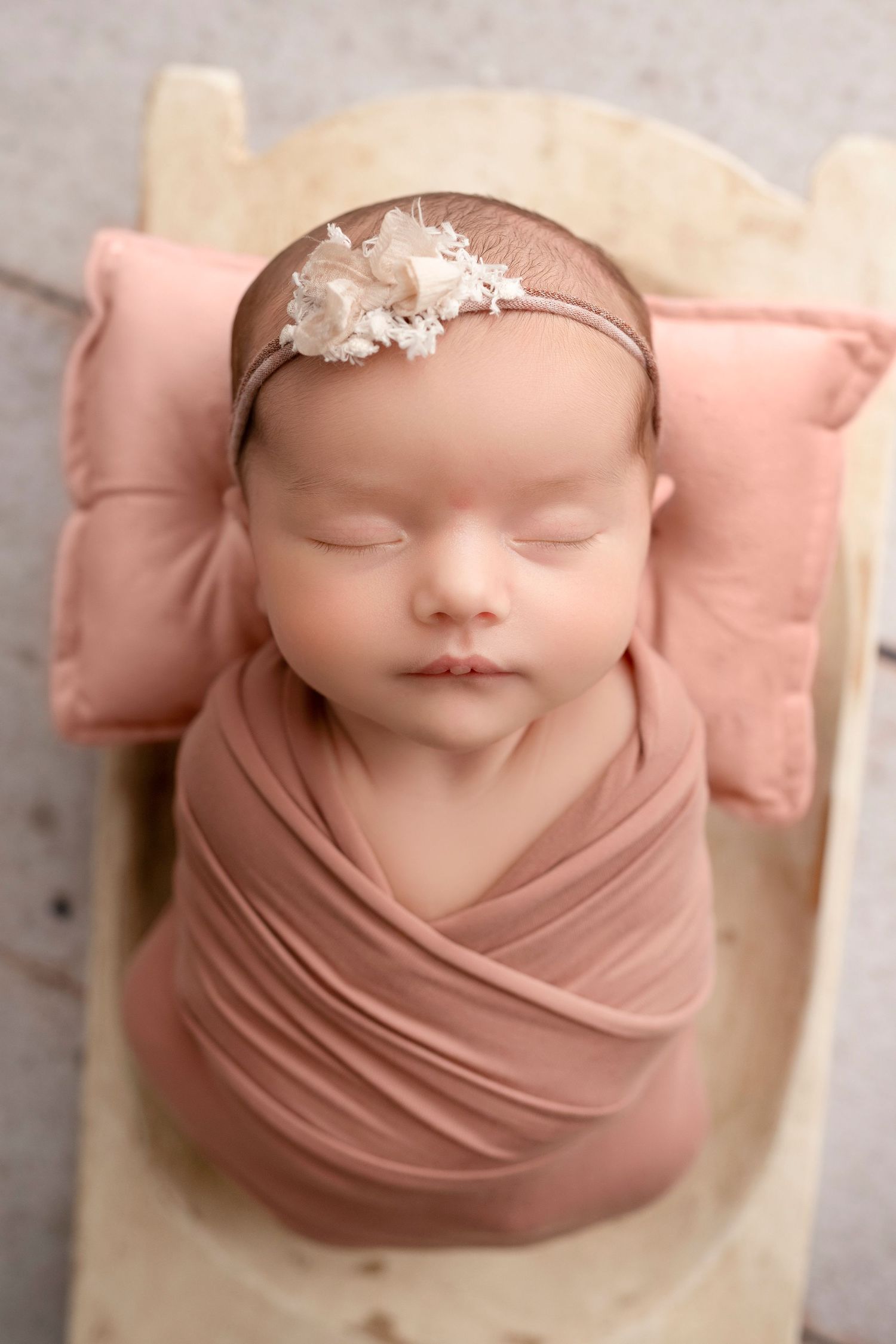 Sleeping newborn wrapped in soft dusty rose fabric with delicate white headpiece against neutral backdrop.