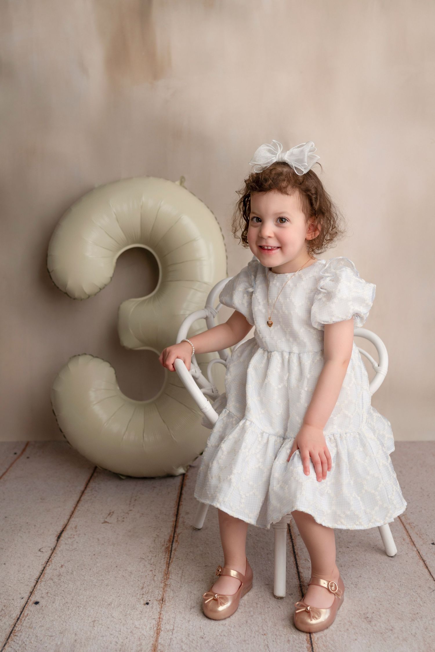 Toddler in white lace dress poses with cream-colored crescent moon prop on wooden floor against beige wall.