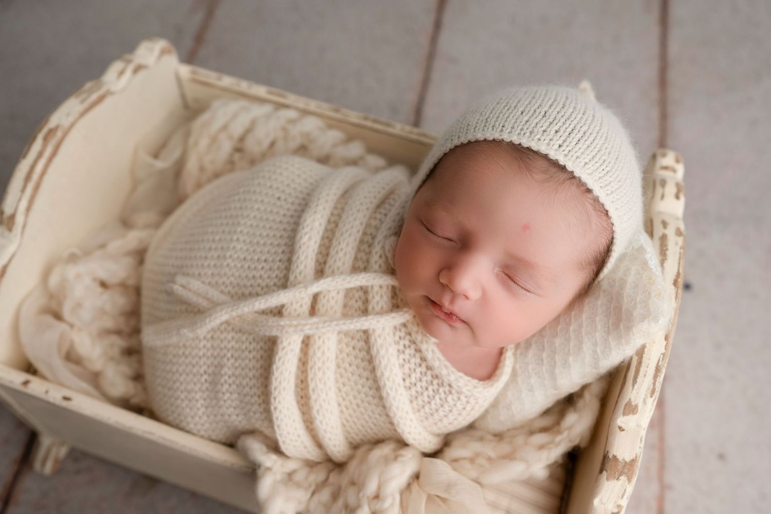 Sleeping newborn baby wearing white knit bonnet resting peacefully in cream-colored basket.